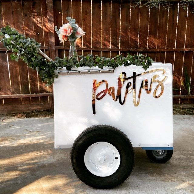 White beverage cart with greenery and flowers, labeled “Paletas,” in front of a wooden fence.