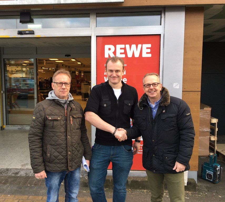 Trzech mężczyzn ściska sobie dłonie przed wejściem do supermarketu REWE.