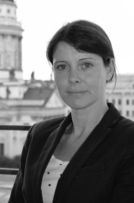 Žena v saku se dívá do kamery, viditelné je městské pozadí.