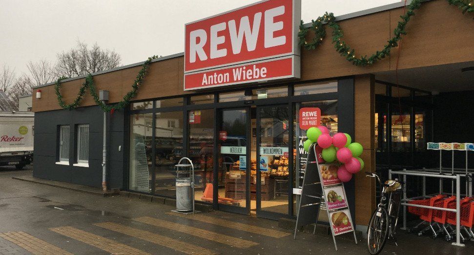Schaufenster eines REWE-Supermarkts mit Luftballons und Grünpflanzen.