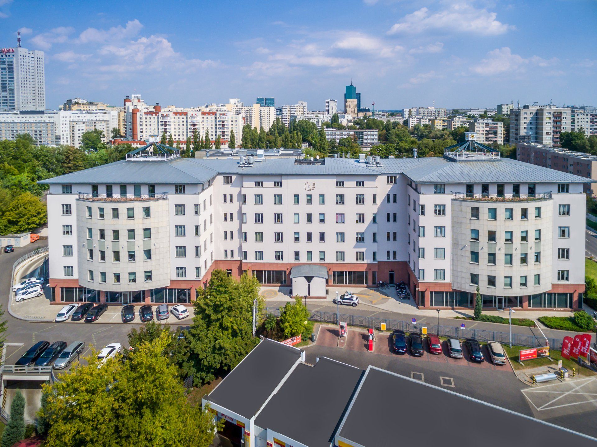 Duży, biały, wielopiętrowy budynek mieszkalny; z przodu drzewa i samochody; w tle panorama miasta.