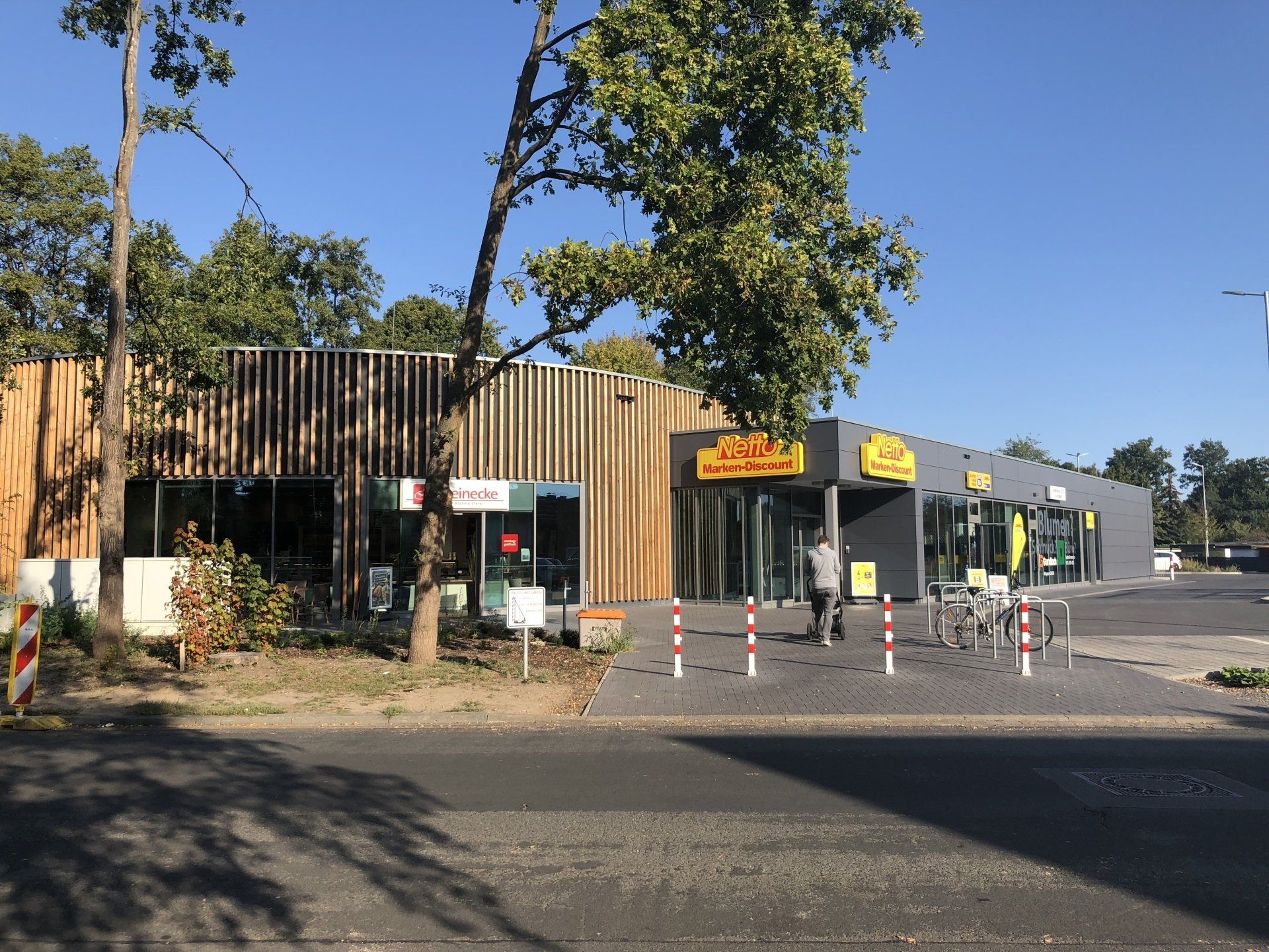 Budovy s obchody, jedna s dřevěnou fasádou. Lidé u vchodů. Jasný, slunečný den.