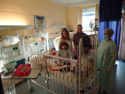 Kinder in Krankenhausbetten, eines mit Engelsflügeln, mit Erwachsenen in einem Krankenhauszimmer.