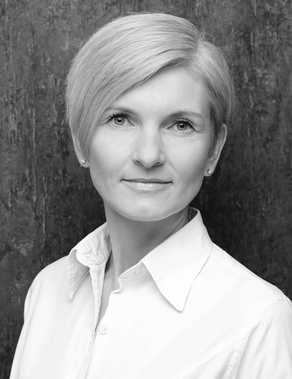 Žena s krátkými blond vlasy v bílé košili s límečkem, s neutrálním výrazem čelí fotoaparátu.