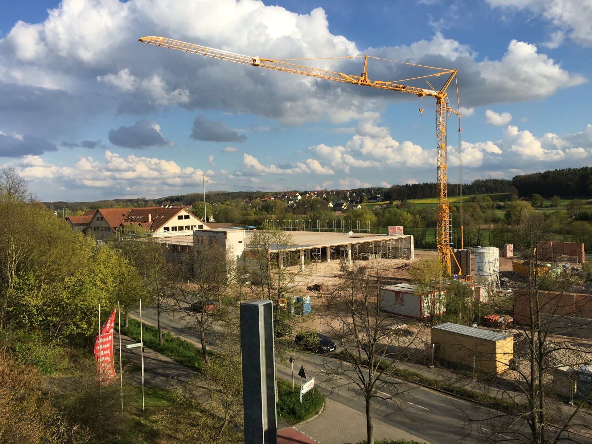 Plac budowy z żółtym dźwigiem, budynkami, drzewami i niebieskim niebem.