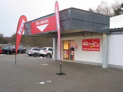 REWE Drive pickup entrance, two teardrop flags, red sign
