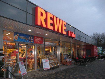 REWE supermarket entrance with red lit sign. Glass doors, food displays, gray building exterior.