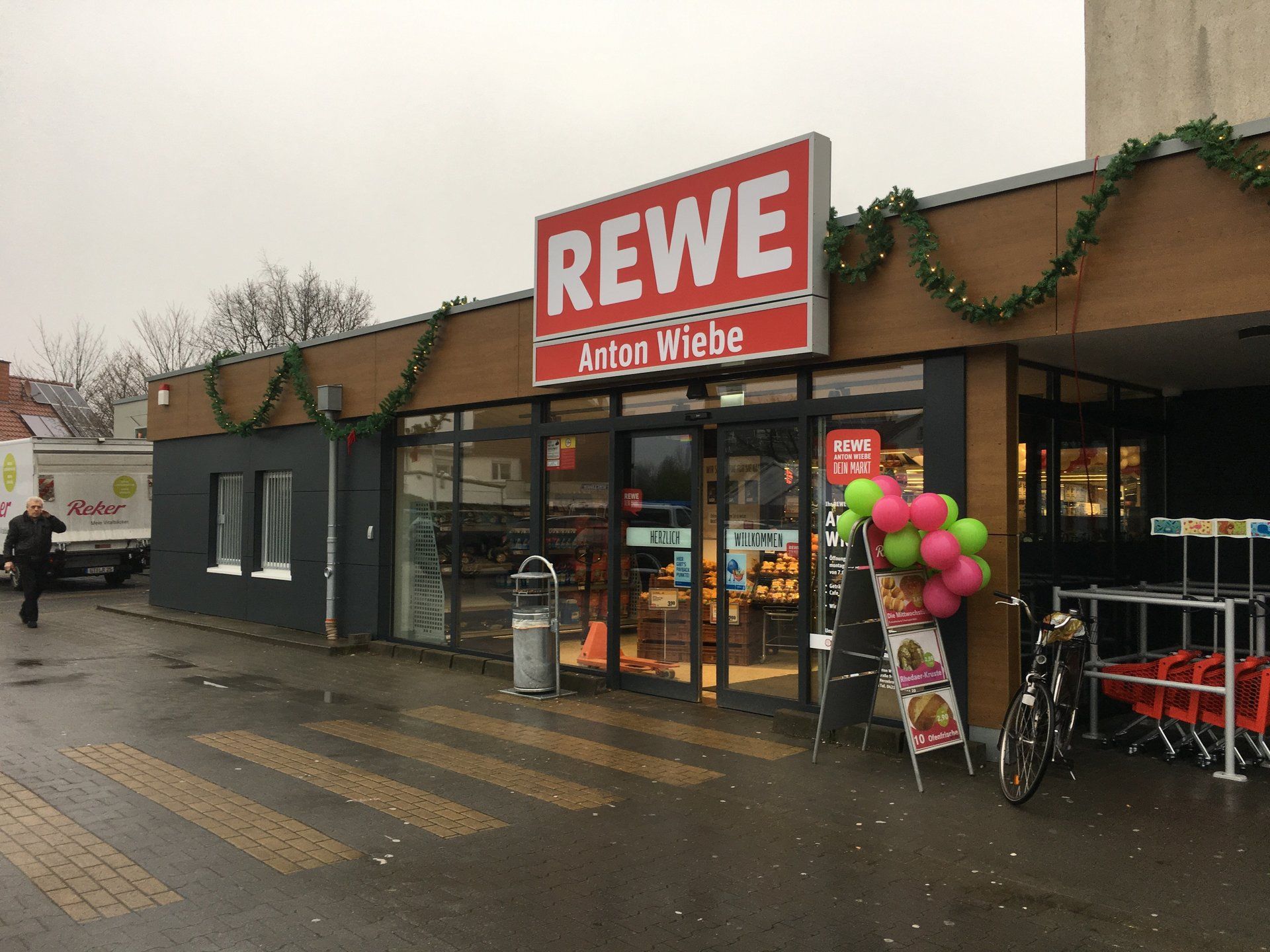 REWEスーパーマーケットの入り口。看板、風船、横断歩道が設置されている。手前を歩いている人。