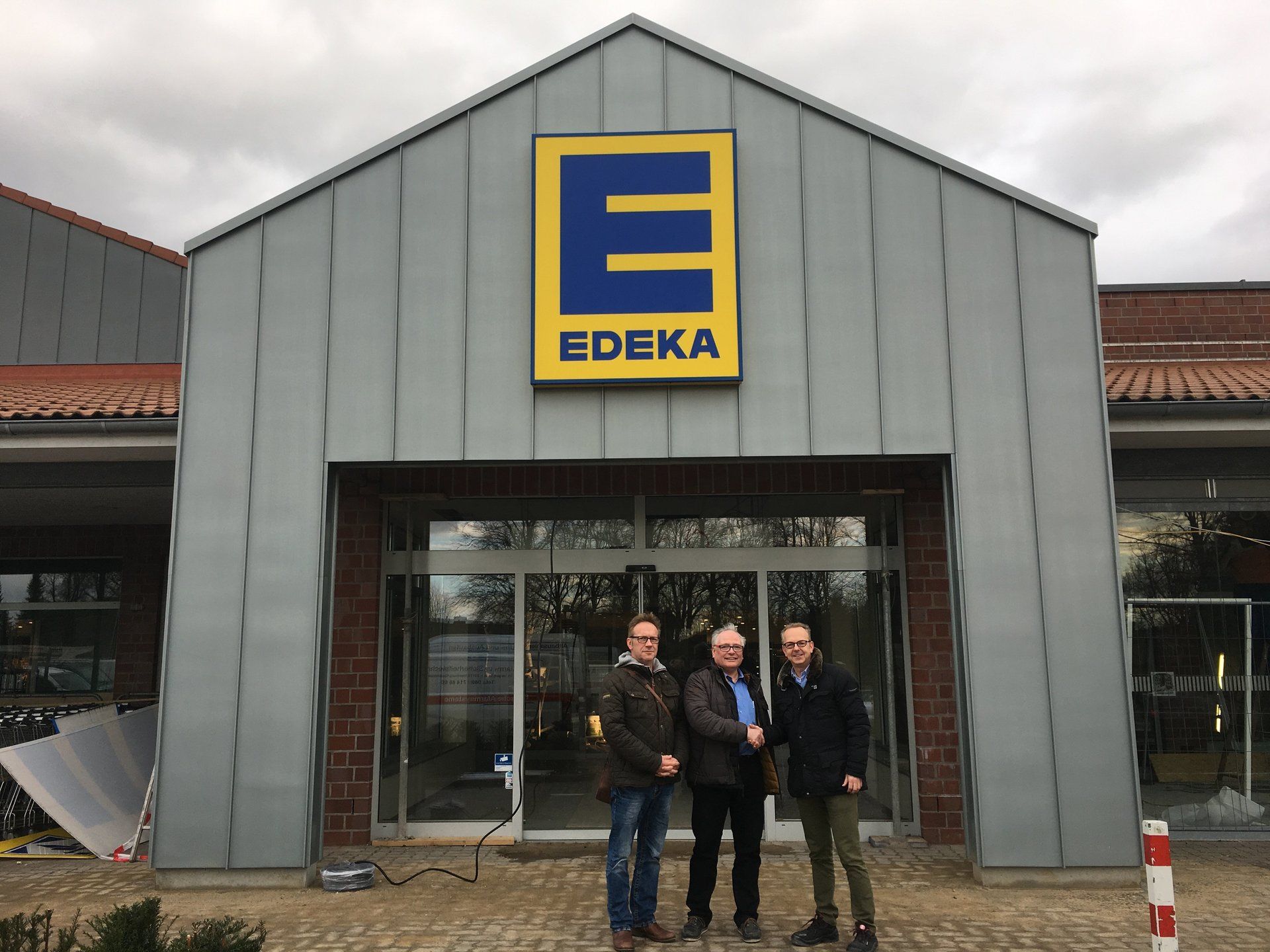 Drei Männer geben sich unter bewölktem Himmel vor dem Eingang eines EDEKA-Lebensmittelgeschäfts die Hand.