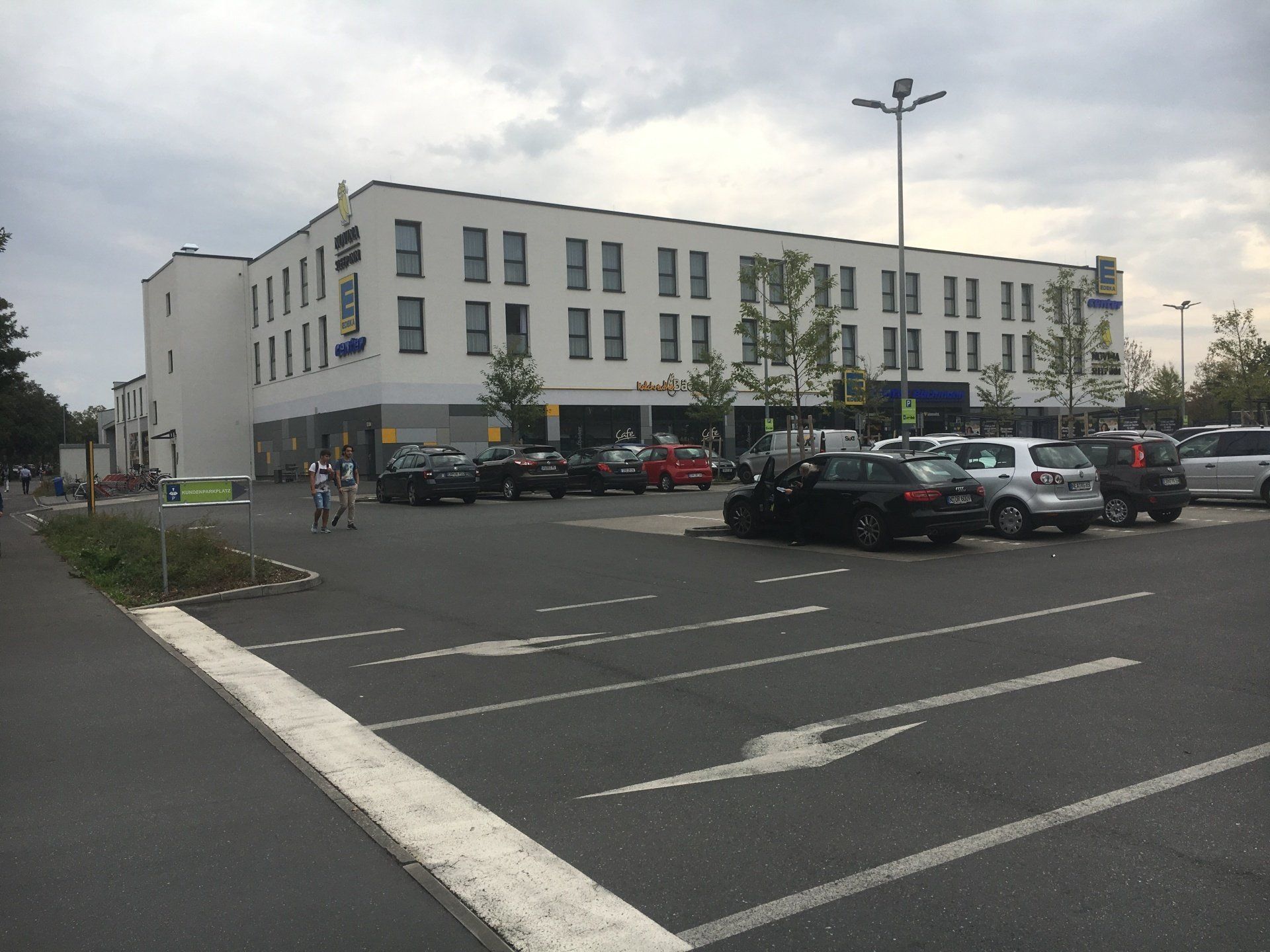Elewacja dwupiętrowego białego budynku z parkingiem przed nim. Zaparkowane samochody. Zachmurzone niebo.
