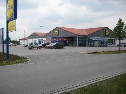 Sklep spożywczy Lidl z samochodami zaparkowanymi przed wejściem w pochmurny dzień. Niebiesko-żółty szyld.