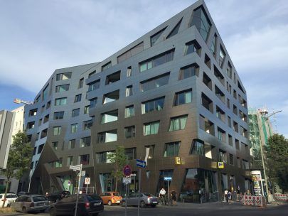 ダークメタルのファサードと大きな窓を備えた、角張ったモダンな建物。駐車車両が並ぶ街の風景。
