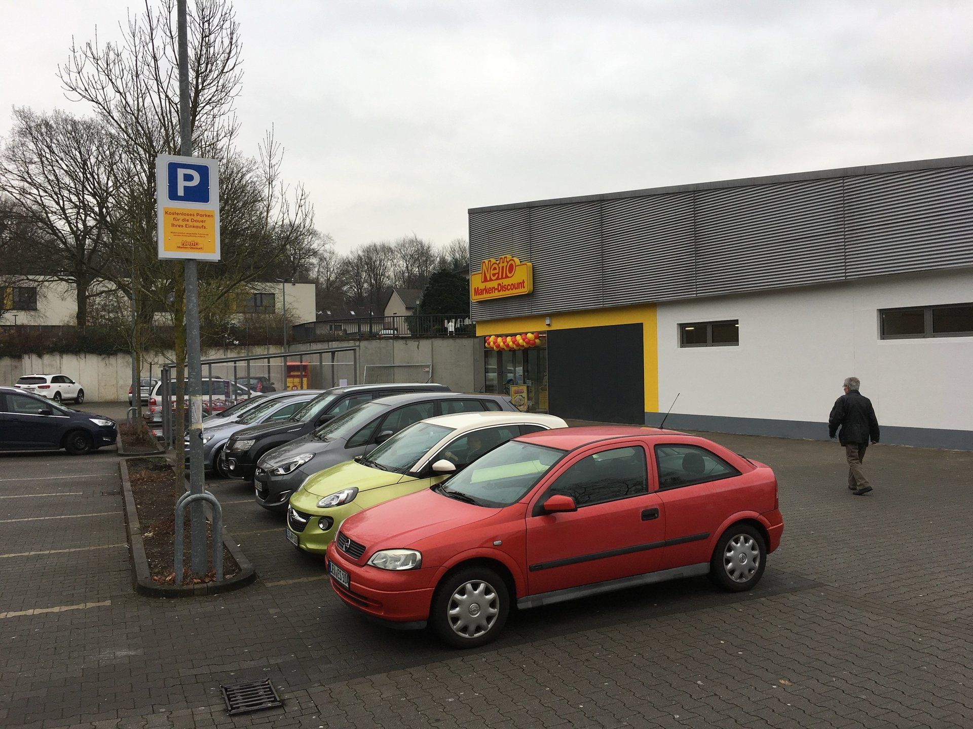 Parking z kilkoma zaparkowanymi samochodami przed supermarketem; przechodzi obok osoba.