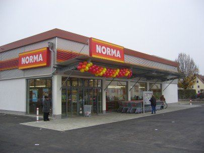 Elewacja supermarketu Norma, czerwone i żółte szyldy, szklane drzwi, kilka osób, pochmurny dzień.
