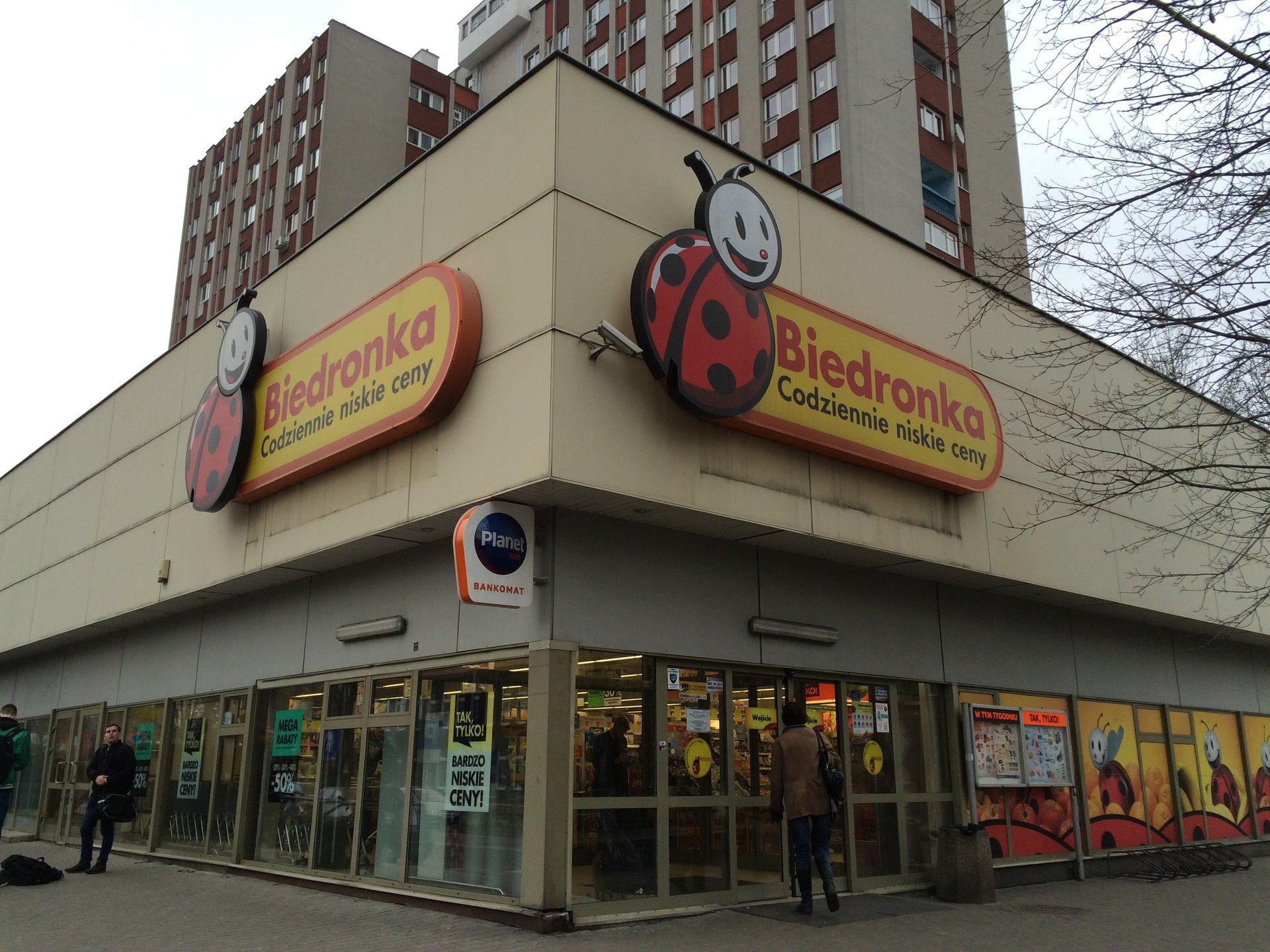 てんとう虫のロゴが入ったビエドロンカ食料品店の外観。人々が店を出入りしている。建物は黄褐色で、背景には高層マンションが見える。