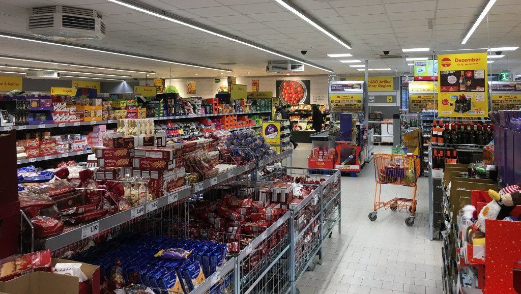 Wnętrze sklepu spożywczego z rzędami półek wypełnionych produktami i kilkoma widocznymi klientami.