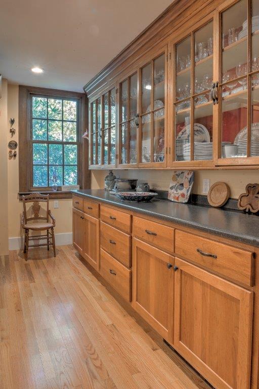 Long wooden kitchen with glass-front cabinets, dark countertops, and a window at the end.