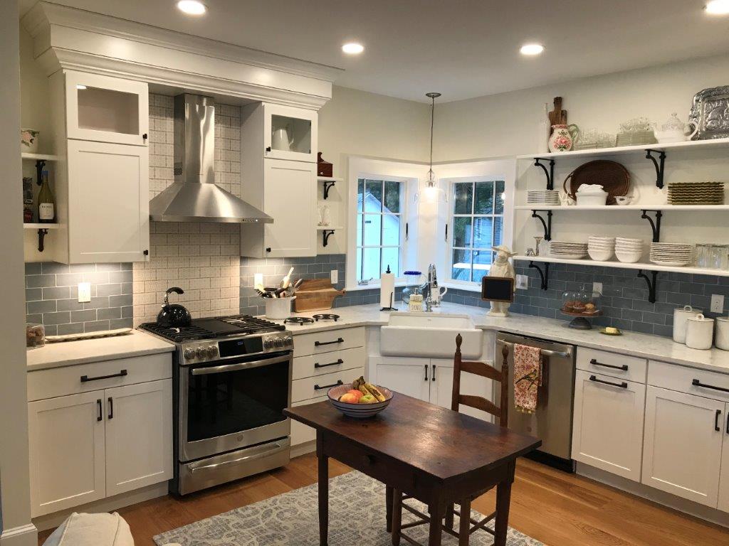 Bright white kitchen with stainless stove, gray tile backsplash, central wooden table, and window seating area