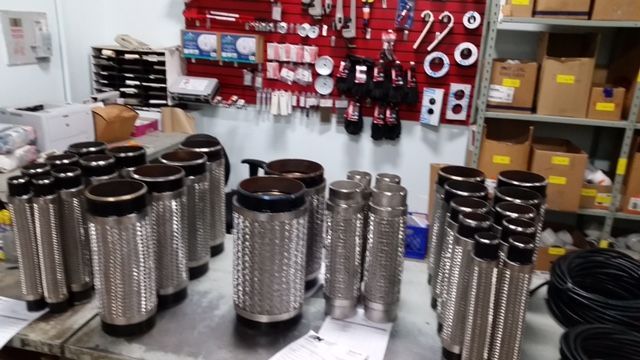 Several stainless steel flexible exhaust pipes on a workbench, with parts and boxes in the background.