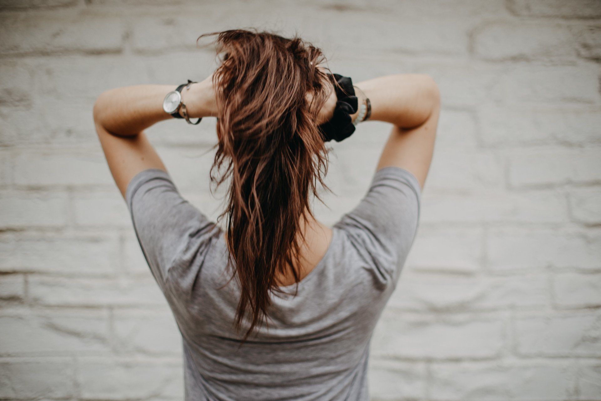Person seen from behind adjusting hair, wearing a gray shirt against a light brick wall.