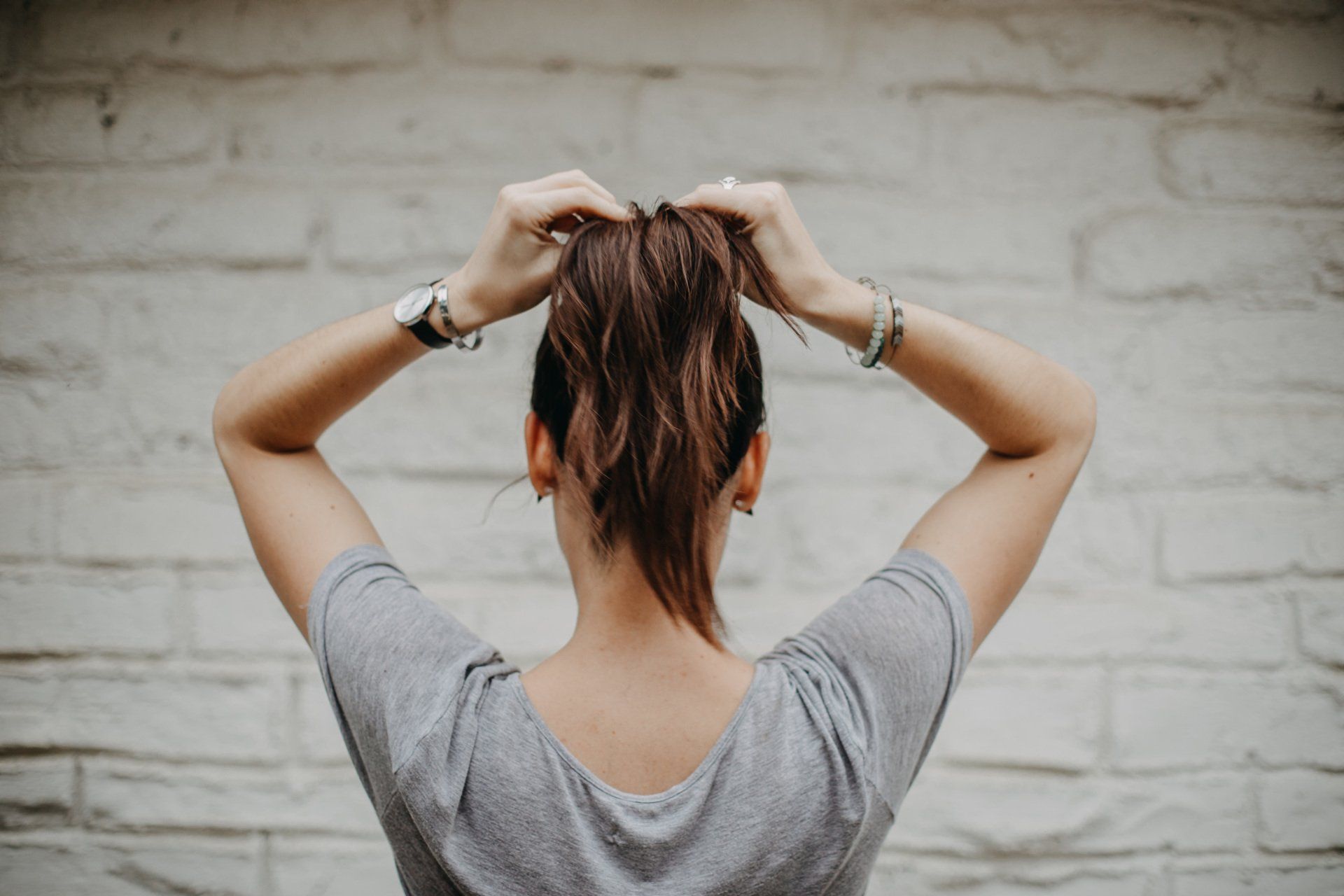 Person seen from behind, tying up long brown hair against a white brick wall.