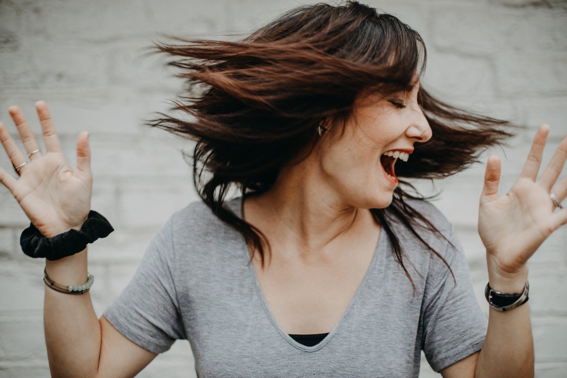 Woman in a gray shirt laughing with hands raised, hair blowing against a plain wall background