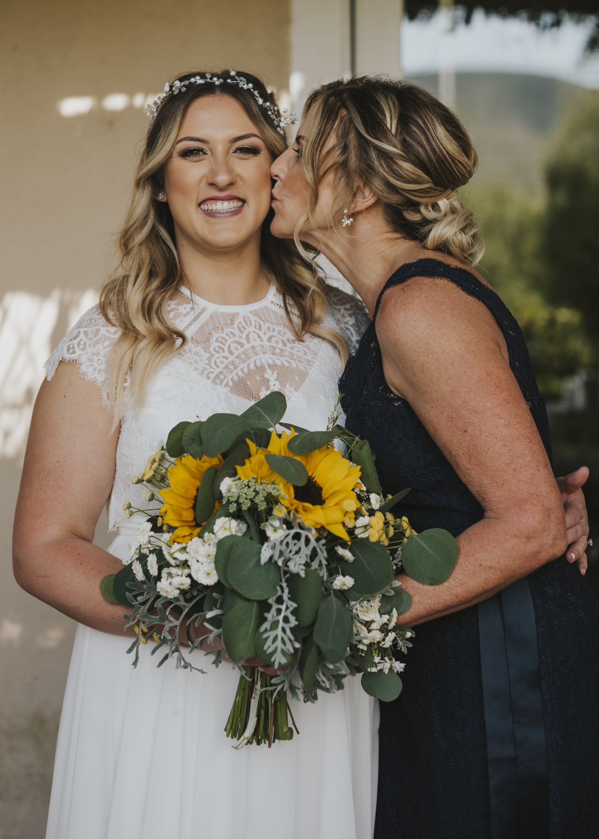 Two women in formal dresses, one kissing the other's cheek while holding a sunflower bouquet.