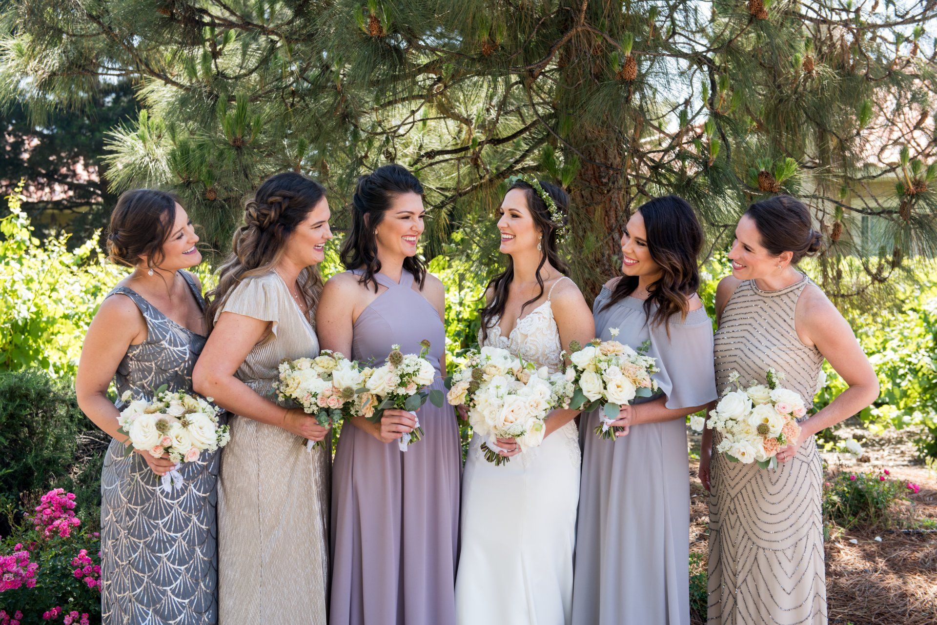 Six women in pastel dresses holding bouquets outdoors under trees at a wedding