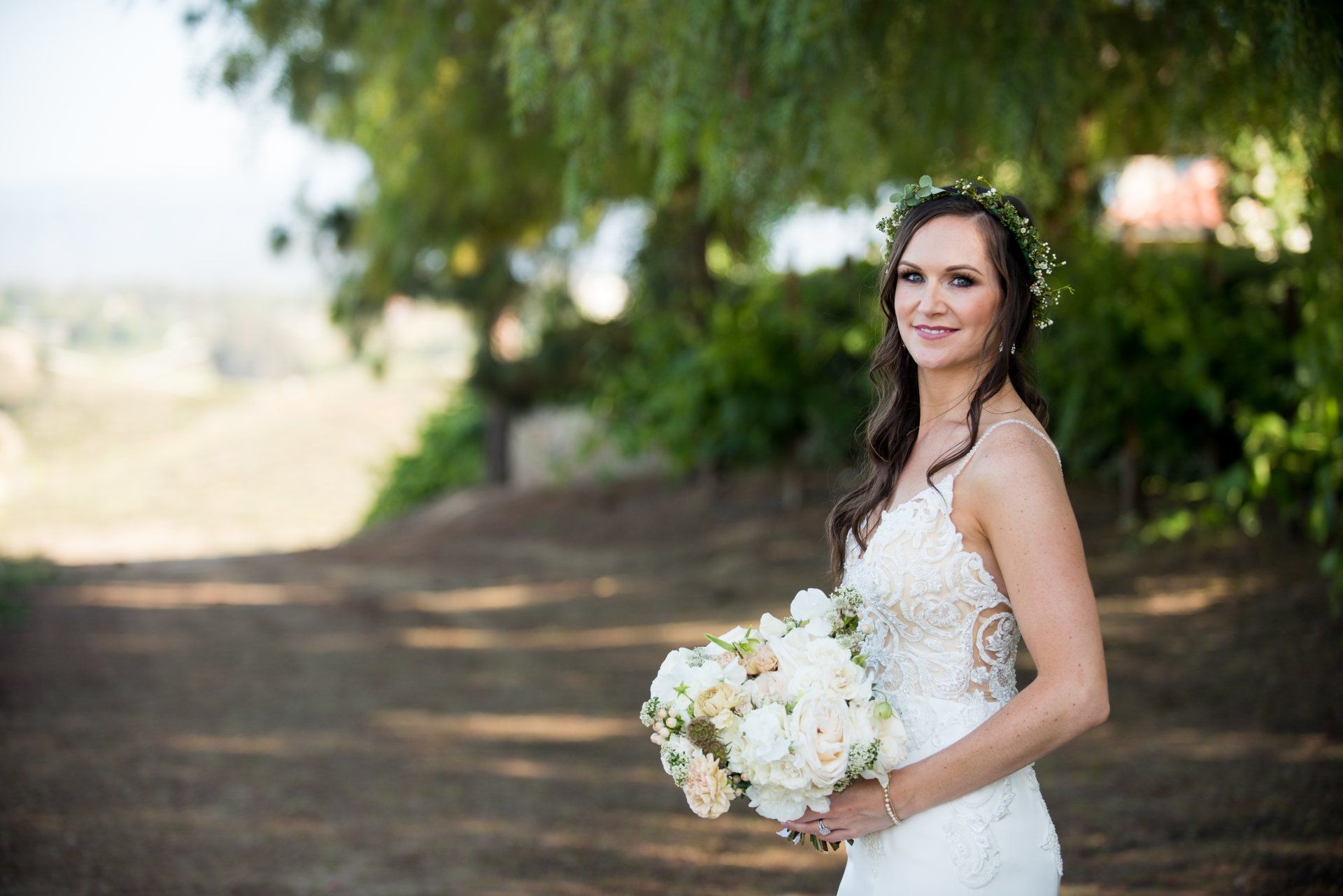 Bride in a white lace dress holding a bouquet outdoors on a shaded path
