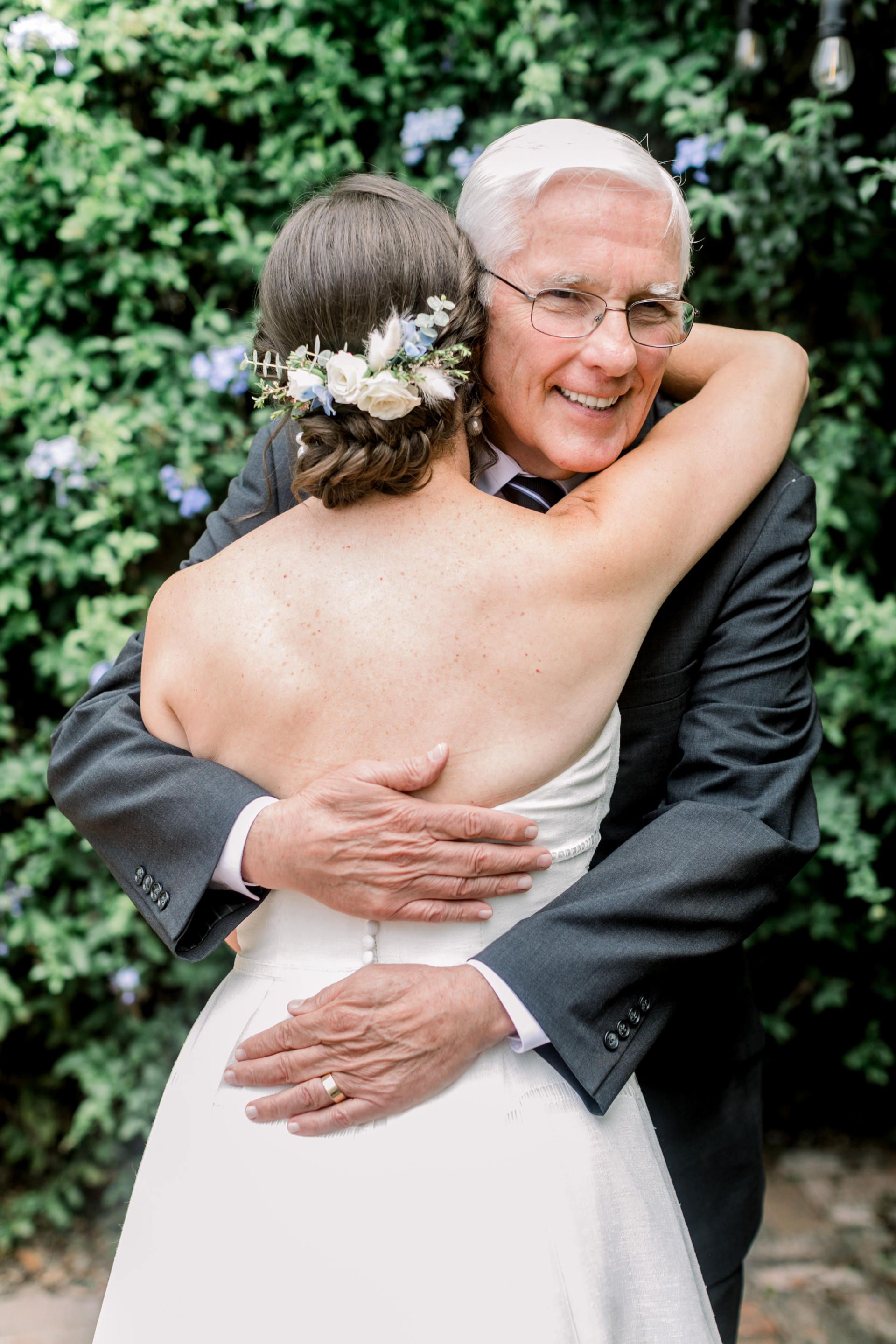 Two people embracing outdoors, with one smiling in a dark suit and the other in a white dress.