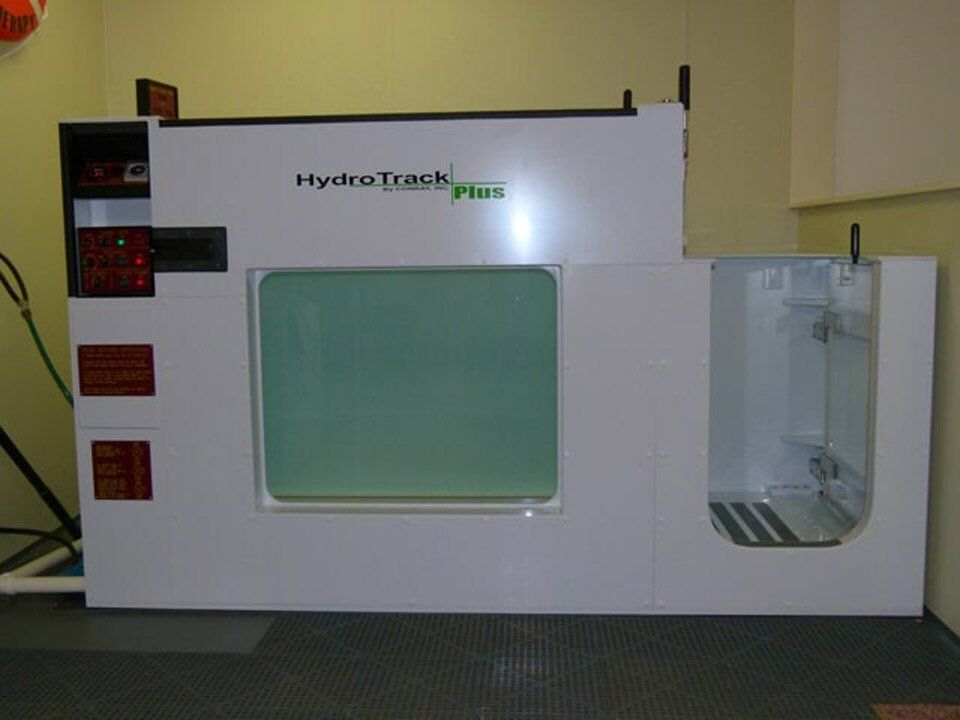 A white HydroTrack underwater treadmill with a viewing window and an entrance door on the right, set in a room.
