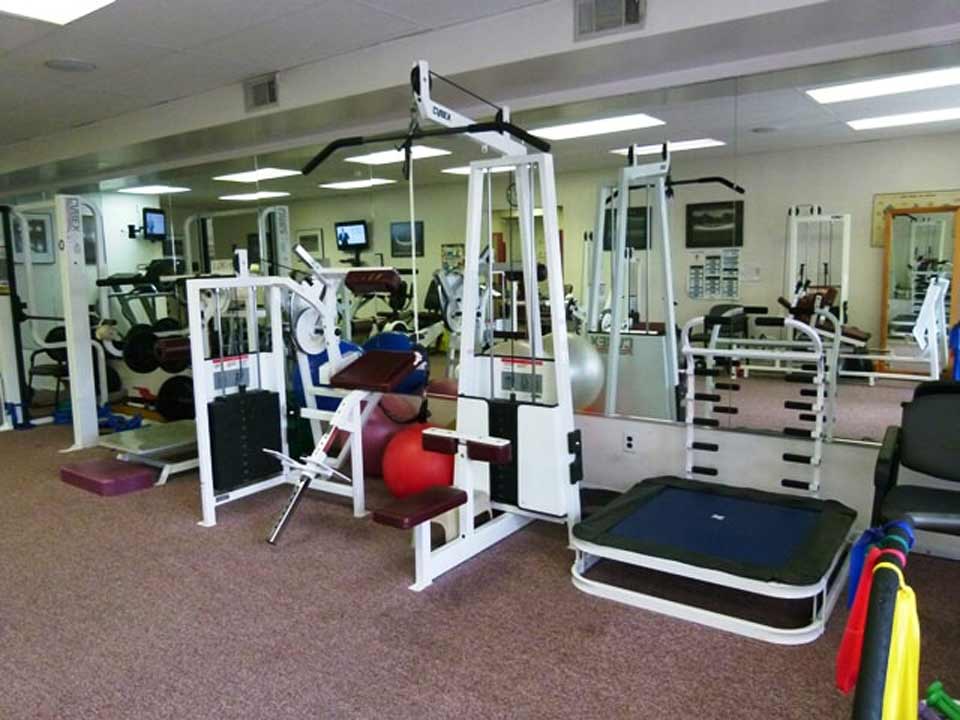 A brightly lit physical therapy gym containing weight machines, a trampoline, and exercise equipment.