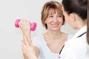 A healthcare professional assists a smiling person with a physical therapy exercise while they hold a pink dumbbell.