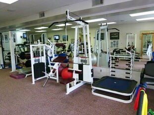 A small gym featuring white strength training machines, a mini trampoline, and mirrors reflecting the workout equipment.