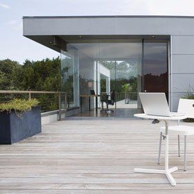 Laptop on a small white table with chair, white footstool holding a glass of red wine, on decking surrounded by steel fencing, outside a one-storey structure with glass walls