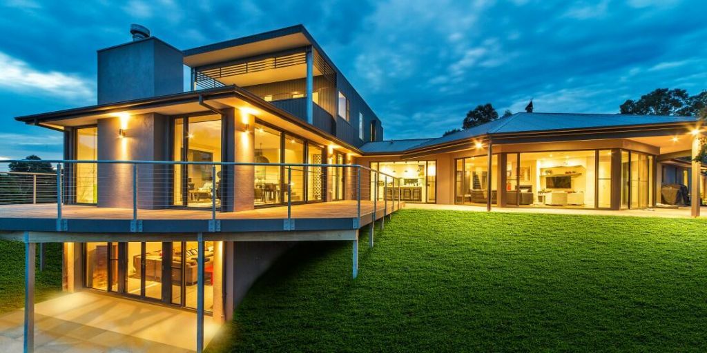 Modern multi-level home with large glass doors, elevated decking and landscaped lawn at dusk.