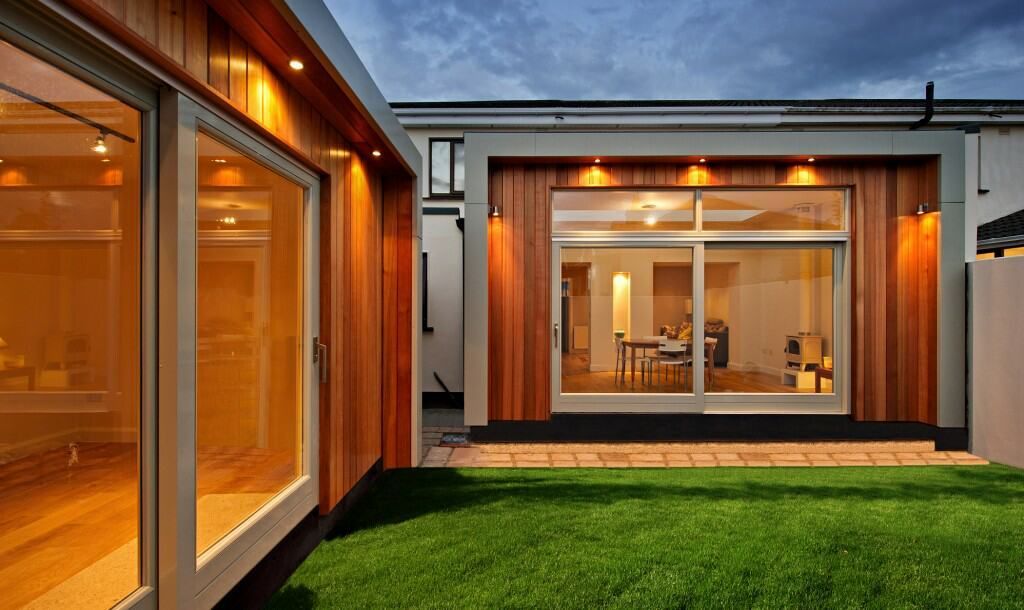 Modern home extension with timber cladding, large windows and a landscaped lawn at dusk.