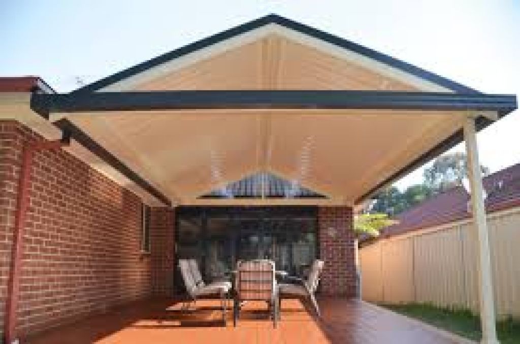 Covered outdoor entertaining area with a pitched roof and dining furniture beside a brick home.