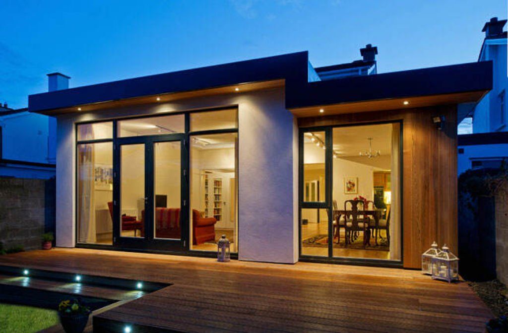 Modern home extension with floor-to-ceiling glass doors, timber cladding and outdoor decking at dusk.