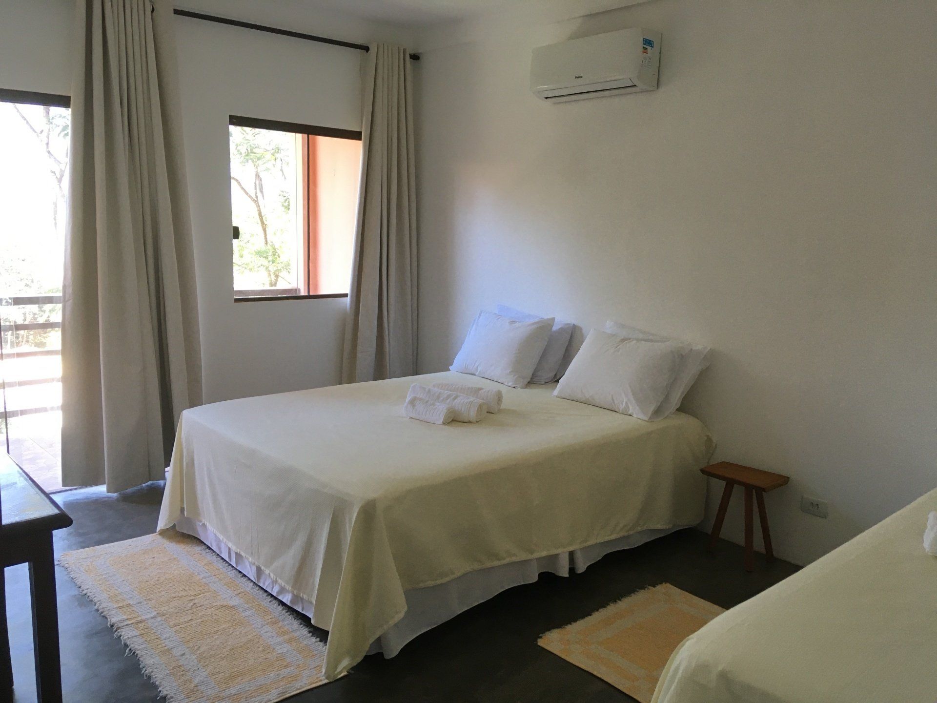 Quarto com duas camas, lençóis cor de creme, paredes brancas e luz natural.