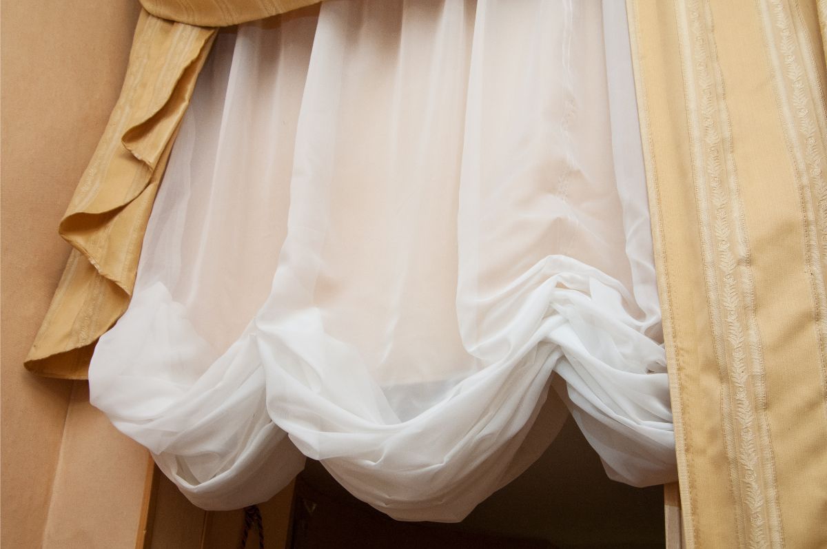 Tessuto bianco trasparente drappeggiato, incorniciato ai lati da tende decorative color beige.