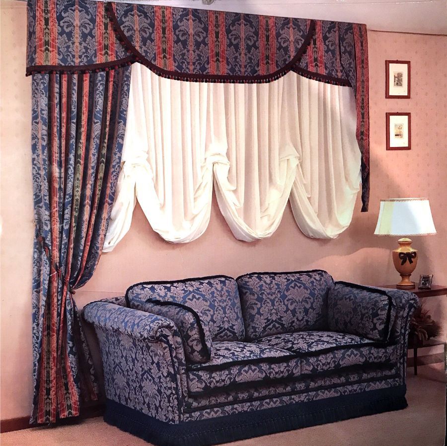 Un divano con motivi blu e rossi è posizionato davanti a una finestra con tende abbinate, una mantovana decorativa e drappeggi bianchi.