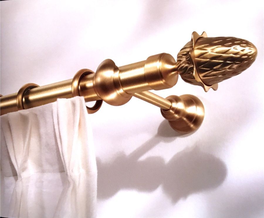 Asta per tende in ottone con un terminale decorativo a forma di pigna, montata a parete con tende bianche appese ad anelli.