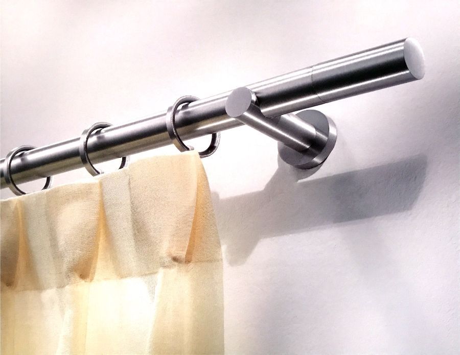 Un'asta per tende in metallo spazzolato montata su una parete bianca, con anelli circolari che sorreggono una tenda in tessuto plissettato di colore chiaro.