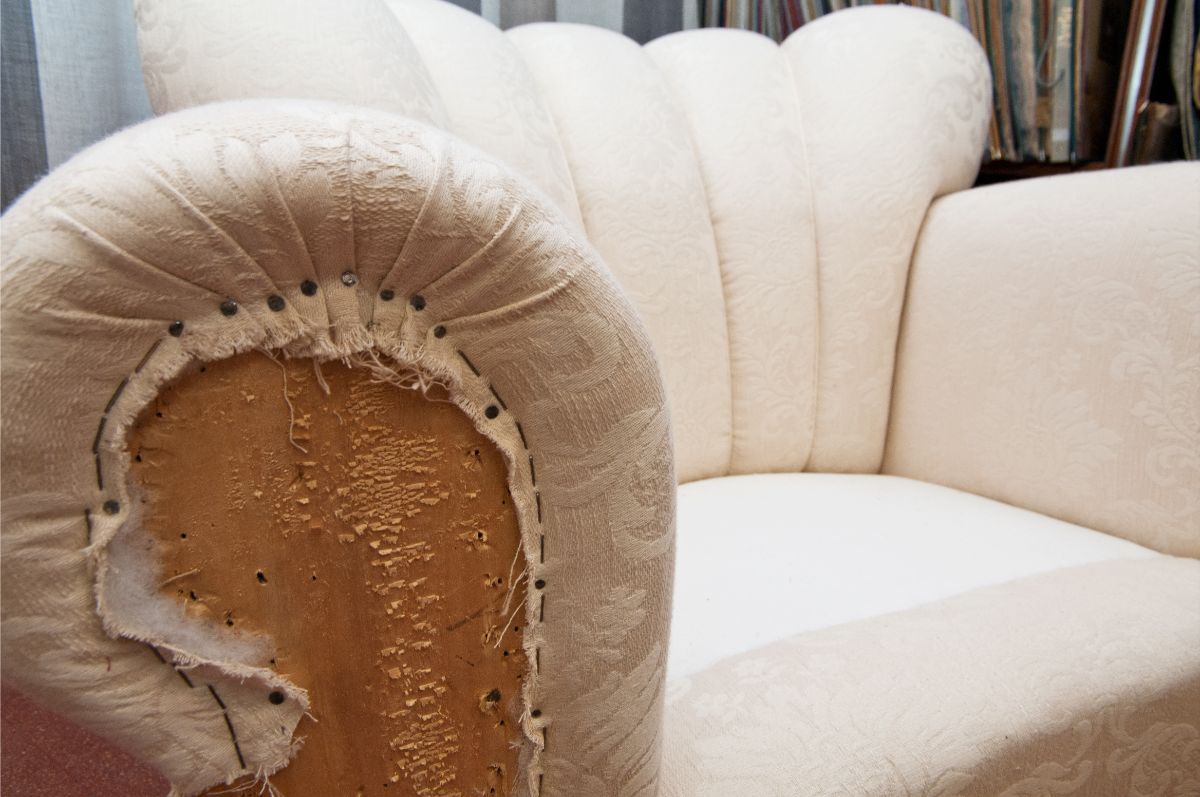 Una poltrona beige con schienale a canali, il cui rivestimento sul bracciolo è danneggiato, lasciando intravedere il tessuto marrone e l'imbottitura sottostanti.