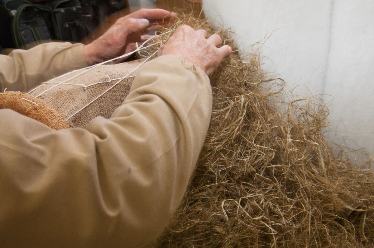Una persona con una camicia color cachi lavora con della tela di iuta su un mucchio di fibre naturali sfuse di colore marrone chiaro.