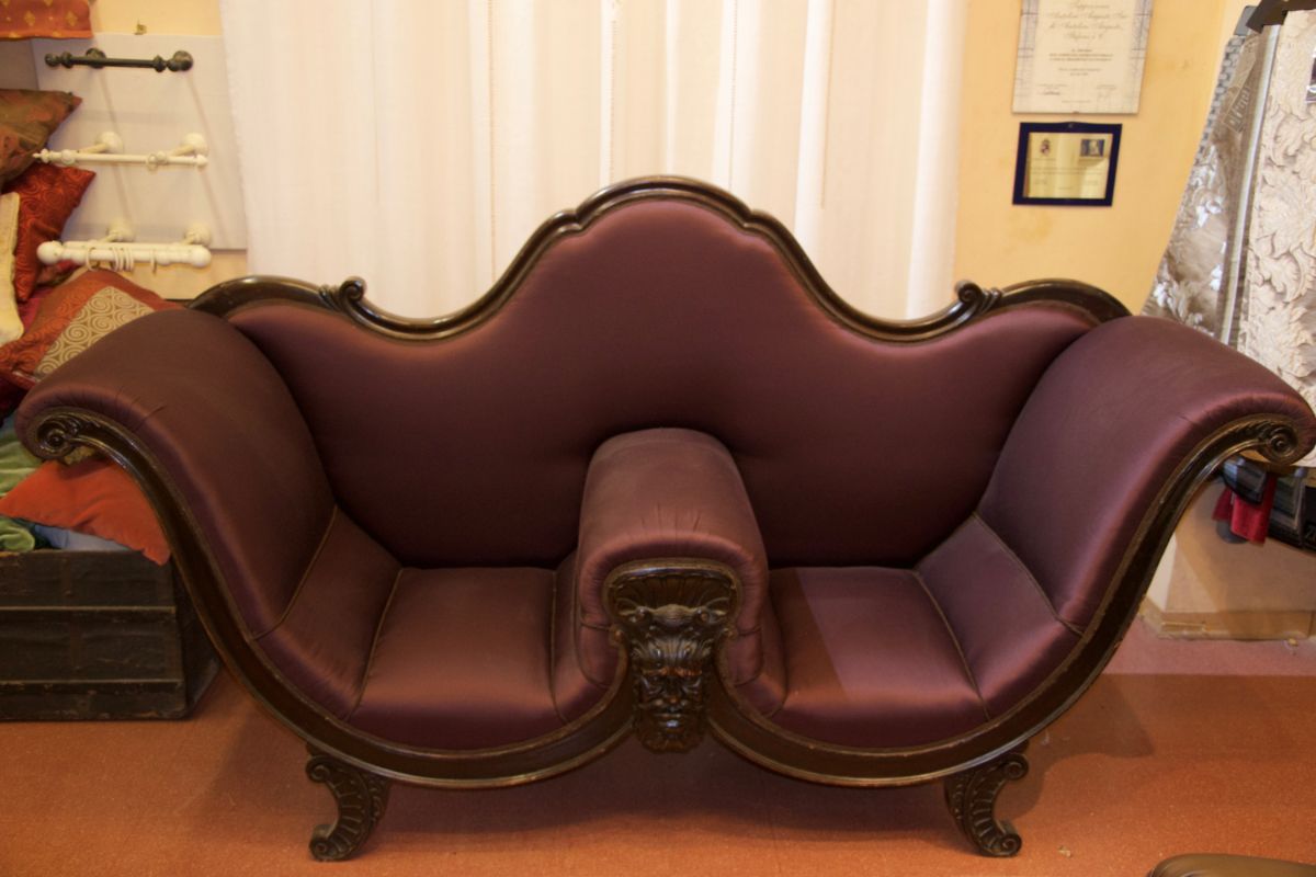 Un divanetto in velluto viola in stile vittoriano con bracciolo centrale e struttura in legno intagliato, collocato in una stanza.