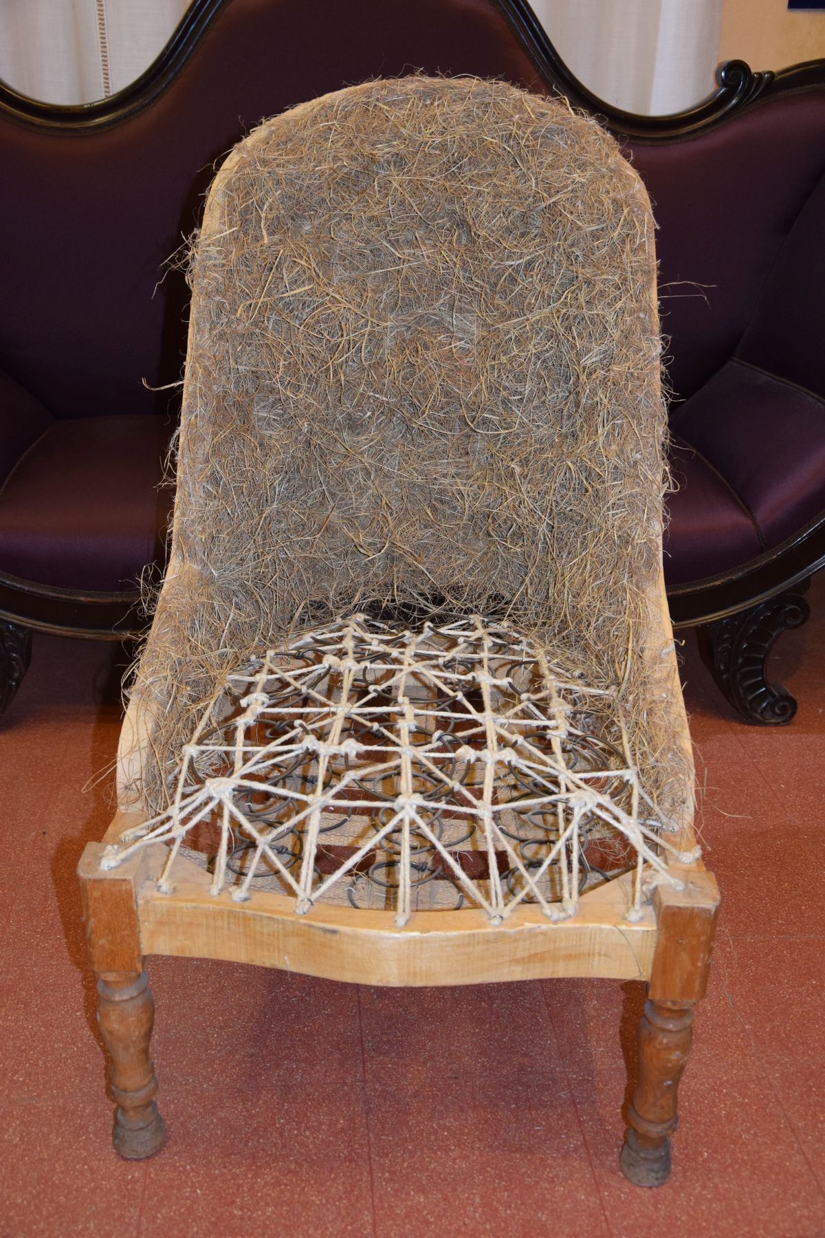 La struttura di una sedia antica mostra le cinghie interne in juta e l'imbottitura in fibra, con un divano viola sullo sfondo sfocato.