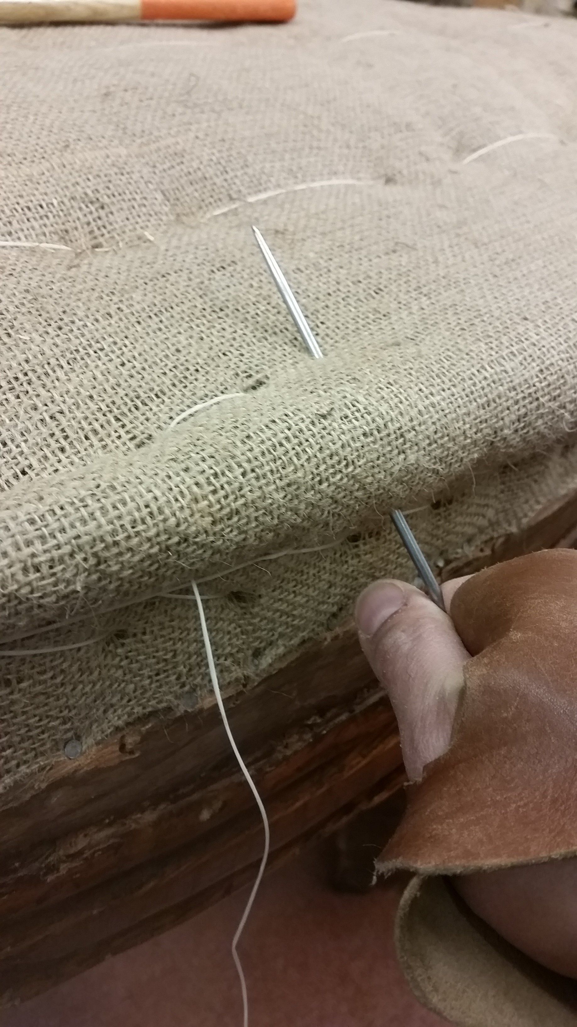 Una mano, protetta da un guanto, spinge un lungo ago da tappezzeria attraverso un tessuto di iuta su un telaio di legno.