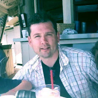 Man sitting at a table with a drink, wearing a plaid shirt in a casual indoor-outdoor setting